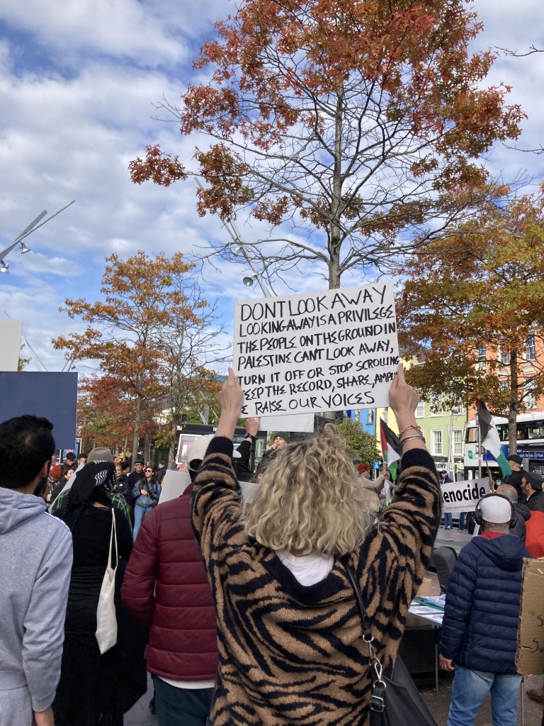 A blonde woman holding up a sign saying: "Don't look away. Looking away is a privilege. The people on the ground in Palestine can't look away, turn it off or stop scrolling. Keep the record, share, amplify & raise our voices."